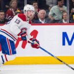 Alexis Lafrenière, wearing a white Montreal Canadiens away jersey with the number 13, skates with the puck on his stick during an ice hockey game. Fans and rink boards with advertisements are visible in the background.