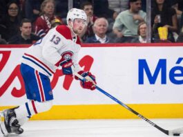 Alexis Lafrenière, wearing a white Montreal Canadiens away jersey with the number 13, skates with the puck on his stick during an ice hockey game. Fans and rink boards with advertisements are visible in the background.