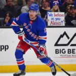 Why Trading Alexis Lafrenière Is Harder Than the New York Rangers Thought New York Rangers forward Alexis Lafrenière, wearing his blue home uniform, skates with the puck during a game. In the background behind the glass, a female fan holds a handwritten sign that reads, "We believe in you Alexis!".