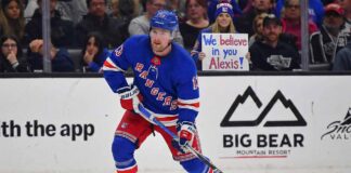 New York Rangers forward Alexis Lafrenière, wearing his blue home uniform, skates with the puck during a game. In the background behind the glass, a female fan holds a handwritten sign that reads, "We believe in you Alexis!".