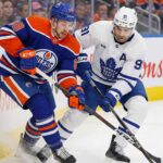 Edmonton Oilers forward Andrew Mangiapane (left) in a blue jersey battles for the puck along the rink boards against Toronto Maple Leafs captain John Tavares in a white away jersey during an NHL game. Their sticks are clashed as they fight for possession.