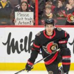 The “Smoke” Around Artem Zub: Why the Senators Facing a Massive Decision Ottawa Senators defenseman Artem Zub (2) kneels on the ice while a female fan behind the glass holds up a white sign with black text that reads, "Please don't trade Artem!" during a game.