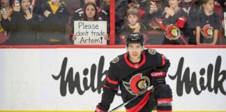 Ottawa Senators defenseman Artem Zub (2) kneels on the ice while a female fan behind the glass holds up a white sign with black text that reads, "Please don't trade Artem!" during a game.