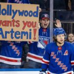 A New York Rangers fan behind the glass holds up a cardboard sign that reads "Hollywood wants you Artemi!" as Rangers forward Artemi Panarin skates on the ice during a game, highlighting trade rumors linking him to the Los Angeles Kings.