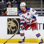 Artemi Panarin, wearing a white New York Rangers away jersey with a blue 'A', is on the ice with his stick during a game. In the background, a fan holds a sign that reads, "Come to Anaheim Artemi!".