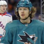 Will the Sharks Land Panarin? The Blockbuster Trade Rumor Heating Up A composite image showing Artemi Panarin in a teal San Jose Sharks uniform and helmet amidst trade speculation, with players wearing white New York Rangers away jerseys visible in the background.