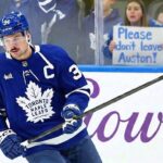 Toronto Maple Leafs captain Auston Matthews skates on the ice during a game, wearing his blue home jersey. Behind the rink glass, a female fan holds up a handwritten white poster board sign with blue text that reads, "Please don't leave Auston!".