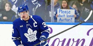 Toronto Maple Leafs captain Auston Matthews skates on the ice during a game, wearing his blue home jersey. Behind the rink glass, a female fan holds up a handwritten white poster board sign with blue text that reads, "Please don't leave Auston!".