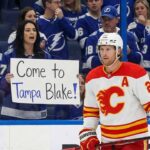 Calgary Flames forward Blake Coleman is on the ice, looking towards a female fan behind the glass. The fan, wearing a Tampa Bay Lightning jersey, holds up a handwritten sign that reads, "Come to Tampa Blake!". Other fans in Lightning gear are visible around her in the stands.