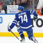 Toronto Maple Leafs forward Bobby McMann (74) kneels on the ice, facing the crowd after a game. Behind the glass in the stands, a female fan with dark hair holds a handmade sign with blue text that reads, "Thanks for the memories Bobby!".