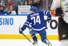 Toronto Maple Leafs forward Bobby McMann (74) kneels on the ice, facing the crowd after a game. Behind the glass in the stands, a female fan with dark hair holds a handmade sign with blue text that reads, "Thanks for the memories Bobby!".