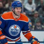 Edmonton Oilers forward Boone Jenner, wearing the blue #38 home jersey, stands on the ice holding his stick during a game, looking towards the play.