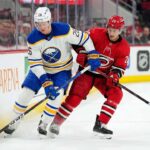 Buffalo Sabres defenseman Rasmus Dahlin (26) in a white away jersey and Carolina Hurricanes forward Sebastian Aho (20) in a red home jersey battle for puck possession along the arena boards during an NHL game at PNC Arena.