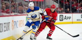 How to Watch Sabres vs. Hurricanes: TV, Stream & Odds (Jan 19, 2026) Buffalo Sabres defenseman Rasmus Dahlin (26) in a white away jersey and Carolina Hurricanes forward Sebastian Aho (20) in a red home jersey battle for puck possession along the arena boards during an NHL game at PNC Arena.