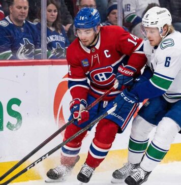 Montreal Canadiens captain Nick Suzuki (in red jersey #14) and Vancouver Canucks alternate captain Brock Boeser (in white jersey #6) battle fiercely for the puck along the boards during an NHL game. Ice spray kicks up around their skates as their sticks clash near the Desjardins advertisement on the boards.