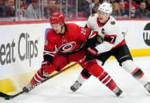 Carolina Hurricanes forward Sebastian Aho (#20) in a red jersey battling with Ottawa Senators captain Brady Tkachuk (#7) in a white jersey for puck possession along the rink boards during an NHL game.