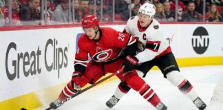 Carolina Hurricanes forward Sebastian Aho (#20) in a red jersey battling with Ottawa Senators captain Brady Tkachuk (#7) in a white jersey for puck possession along the rink boards during an NHL game.