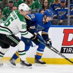 Dallas Stars forward Mikko Rantanen (96), wearing a white away jersey, battles for puck possession along the boards against St. Louis Blues forward Brayden Schenn (10), wearing a blue home jersey.