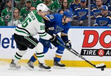 Dallas Stars forward Mikko Rantanen (96), wearing a white away jersey, battles for puck possession along the boards against St. Louis Blues forward Brayden Schenn (10), wearing a blue home jersey.