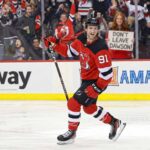 New Jersey Devils forward Dawson Mercer smiles on the ice while a fan behind the glass holds up a handwritten "DON'T LEAVE DAWSON!" sign during a game.