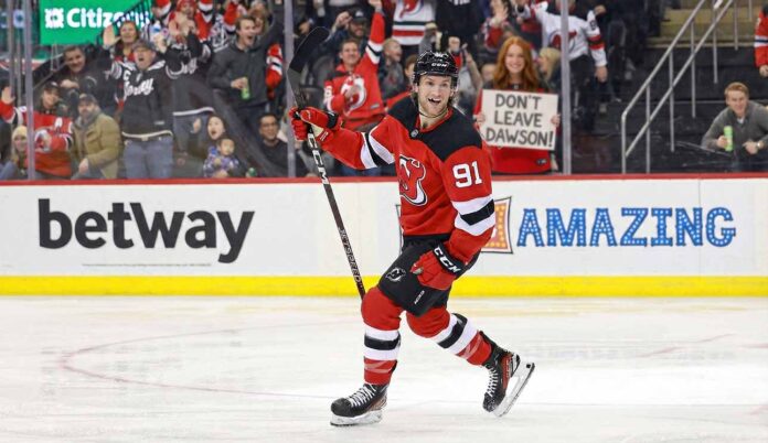 New Jersey Devils forward Dawson Mercer smiles on the ice while a fan behind the glass holds up a handwritten "DON'T LEAVE DAWSON!" sign during a game.