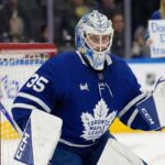 Toronto Maple Leafs goaltender Dennis Hildeby, wearing number 35, stands ready in his crease during a game. Behind the protective glass in the blurred background crowd, a blonde female fan holds up a white handmade sign with blue lettering that reads, "Don't leave Dennis, trade Stolarz."