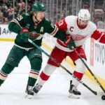 Minnesota Wild's Kirill Kaprizov (#97 in green jersey) and Detroit Red Wings' Dylan Larkin (#71 in white jersey) battle for possession of the puck against the rink boards during an NHL game.