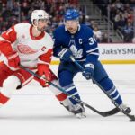 Auston Matthews in a blue Toronto Maple Leafs jersey with the Captain's C battling Dylan Larkin in a white Detroit Red Wings jersey for puck possession during an NHL game.
