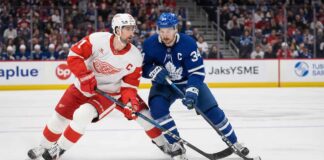 Auston Matthews in a blue Toronto Maple Leafs jersey with the Captain's C battling Dylan Larkin in a white Detroit Red Wings jersey for puck possession during an NHL game.