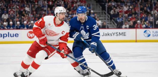 Will the Wings Stun Toronto? How to Watch Red Wings vs. Maple Leafs Live Tonight Auston Matthews in a blue Toronto Maple Leafs jersey with the Captain's C battling Dylan Larkin in a white Detroit Red Wings jersey for puck possession during an NHL game.