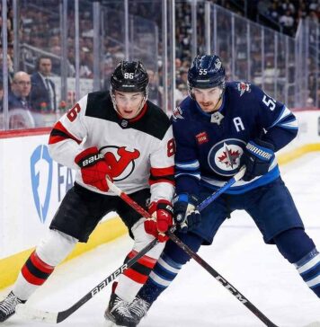 New Jersey Devils center Jack Hughes (white jersey) battles for the puck against Winnipeg Jets center Mark Scheifele (blue jersey) during an NHL game at Canada Life Centre.