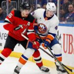 New Jersey Devils forward Jack Hughes (86), wearing a red jersey with an alternate captain 'A' patch, battles for possession of the puck against New York Islanders defenseman Matthew Schaefer (48), wearing a white jersey, along the rink boards.