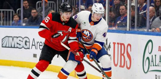 New Jersey Devils forward Jack Hughes (86), wearing a red jersey with an alternate captain 'A' patch, battles for possession of the puck against New York Islanders defenseman Matthew Schaefer (48), wearing a white jersey, along the rink boards.