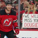 New Jersey Devils defenseman Dougie Hamilton smiles broadly while wearing his red home jersey and gear on the ice. Behind the arena glass in the stands, a female fan with blonde hair and a Devils cap holds up a handwritten white sign with black and red text that reads, "WE WILL MISS YOU DOUGIE!".