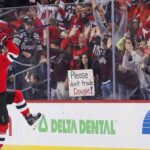 New Jersey Devils defenseman Dougie Hamilton (#7) in a red home jersey celebrates against the glass, lifting his leg and stick. Behind the glass, a crowd of cheering fans watches, including a woman with dark hair prominently holding a handmade white sign that reads, "Please don't trade Dougie!".