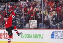 New Jersey Devils defenseman Dougie Hamilton (#7) in a red home jersey celebrates against the glass, lifting his leg and stick. Behind the glass, a crowd of cheering fans watches, including a woman with dark hair prominently holding a handmade white sign that reads, "Please don't trade Dougie!".