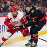 Detroit Red Wings captain Dylan Larkin (in white away jersey) and Ottawa Senators captain Brady Tkachuk (in black home jersey) intensely battling for the puck along the arena boards during an NHL game.