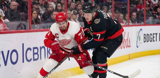Detroit Red Wings captain Dylan Larkin (in white away jersey) and Ottawa Senators captain Brady Tkachuk (in black home jersey) intensely battling for the puck along the arena boards during an NHL game.