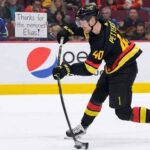Elias Pettersson (#40) of the Vancouver Canucks, wearing his black "Flying V" jersey, winds up for a slapshot on the ice. Behind him, a female fan with dark hair stands in the front row behind the glass, holding a white sign with handwritten black text that reads, "Thanks for the memories Elias!". Other fans fill the red seats in the background.