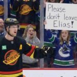 Vancouver Canucks alternate captain Elias Pettersson stands on the ice in his black alternate jersey, while a female fan behind the glass holds up a handwritten white sign that reads, "Please don't leave Elias!".