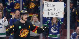 Vancouver Canucks alternate captain Elias Pettersson stands on the ice in his black alternate jersey, while a female fan behind the glass holds up a handwritten white sign that reads, "Please don't leave Elias!".