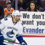 Is the Door Closed? The Real Story Behind the Evander Kane to Oilers Rumors Vancouver Canucks player Evander Kane, wearing a white number 91 away jersey, skates along the boards on the ice. Behind the protective glass, a female fan with a stern expression holds up a white sign with bold text that reads, "We don't want you Evander!" She is surrounded by other spectators in the stands, many wearing Edmonton Oilers jerseys.