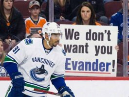 Is the Door Closed? The Real Story Behind the Evander Kane to Oilers Rumors Vancouver Canucks player Evander Kane, wearing a white number 91 away jersey, skates along the boards on the ice. Behind the protective glass, a female fan with a stern expression holds up a white sign with bold text that reads, "We don't want you Evander!" She is surrounded by other spectators in the stands, many wearing Edmonton Oilers jerseys.