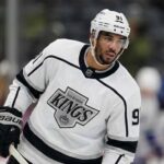 Evander Kane standing on an ice hockey rink, wearing a white Los Angeles Kings away jersey with the Kings crown logo and number 91 on the sleeve, holding his stick and looking to the side.