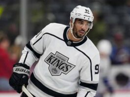 Evander Kane standing on an ice hockey rink, wearing a white Los Angeles Kings away jersey with the Kings crown logo and number 91 on the sleeve, holding his stick and looking to the side.