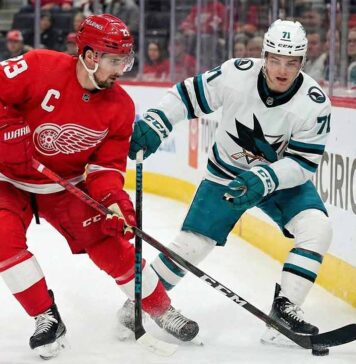 Detroit Red Wings captain Dylan Larkin and San Jose Sharks Macklin Celebrini battle with their sticks for the puck along the arena boards during an NHL game.