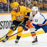 Nashville Predators captain Roman Josi (#59) in a yellow jersey and New York Islanders forward Mathew Barzal (#13) in a white jersey battle for the puck along the boards during an NHL game at Bridgestone Arena.
