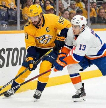 Nashville Predators captain Roman Josi (#59) in a yellow jersey and New York Islanders forward Mathew Barzal (#13) in a white jersey battle for the puck along the boards during an NHL game at Bridgestone Arena.