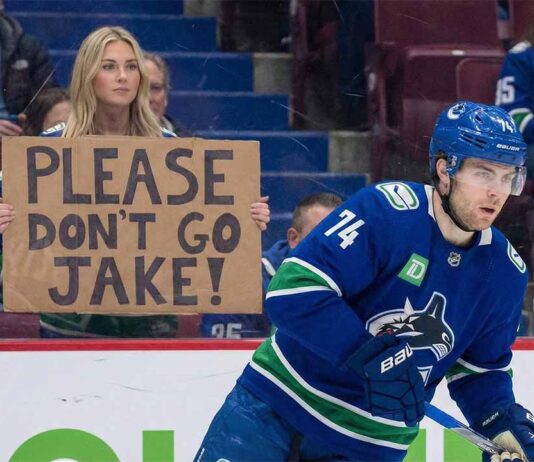 Vancouver Canucks forward Jake DeBrusk (74) skates on the ice during a game, while a female fan behind the protective glass holds up a handwritten cardboard sign that reads, "PLEASE DON'T GO JAKE!" amidst ongoing trade speculation.