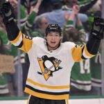 ason Robertson, wearing a white Pittsburgh Penguins away uniform, smiles broadly with his arms raised in celebration on the ice. Behind the rink glass, a crowd of fans, many wearing green, cheers. On the left, a blurred female fan holds a handwritten cardboard sign that reads, "Don't leave Jason!"